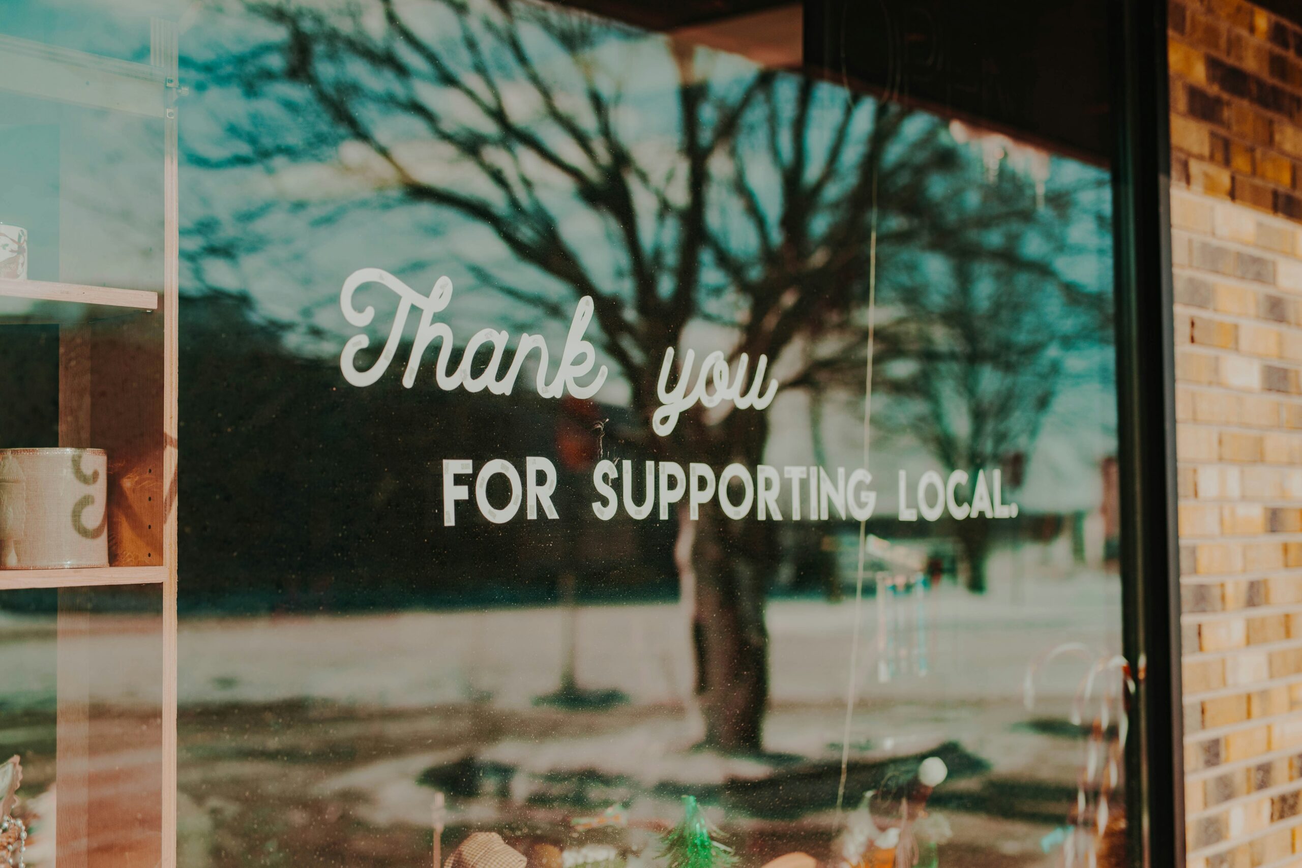Hand-lettered "Thank you for supporting local" sign in a small business storefront window, with a tree-lined street reflected in the glass